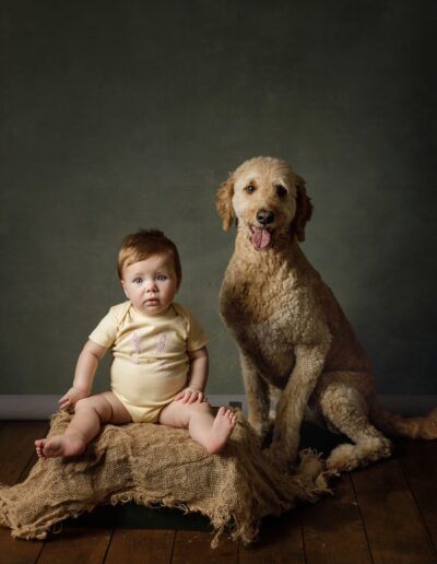 Baby sitting beside a goldendoodle on a hessian-covered crate, family pet photoshoot in Barking