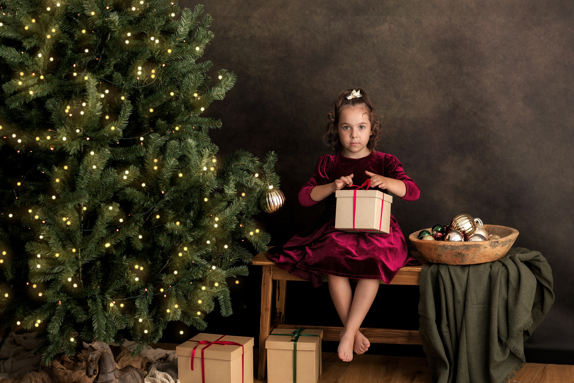 Little girl in a red velvet dress opening a gift by the Christmas tree - EvaGud Photography Christmas Mini Sessions 2026