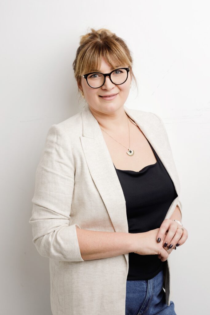 Business headshot of a woman in a cream blazer and black top, hands together, against a bright white backdrop
