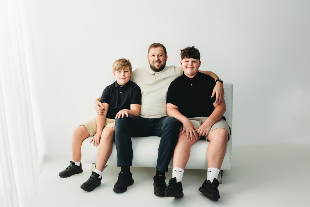 Dad and his two sons sitting side by side on a white sofa in a relaxed studio portrait
