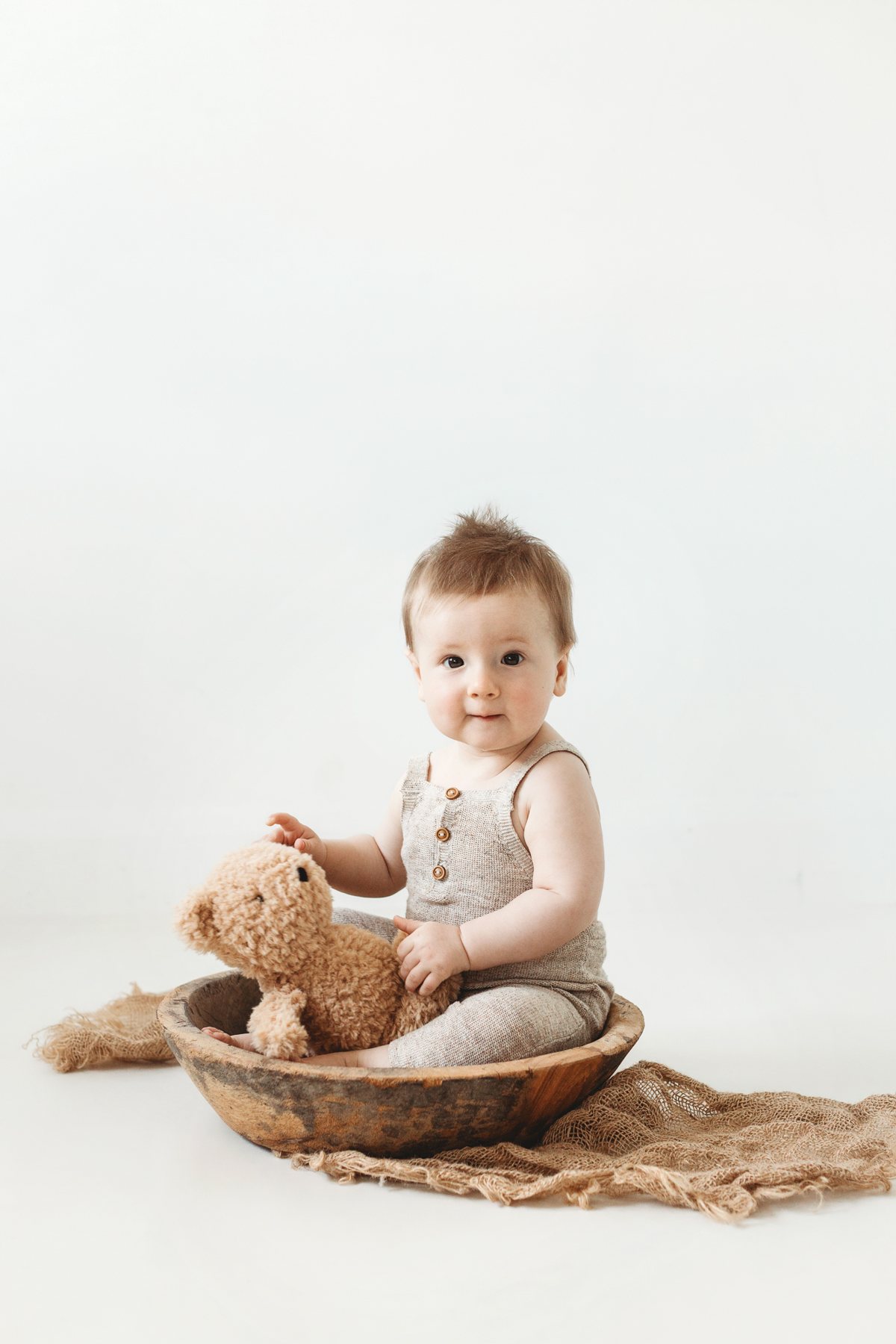 Baby sitting in a wooden bowl with a teddy bear at a Milestone Mini Session