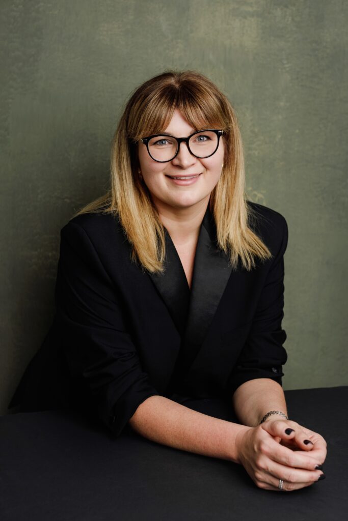 Professional headshot of a smiling woman with glasses seated at a table with her hands clasped, green textured backdrop