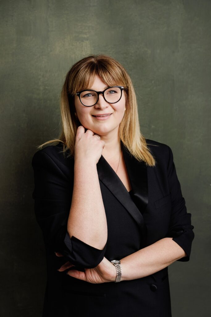 Professional headshot of a woman with glasses resting her chin on her hand, black blazer, green textured backdrop