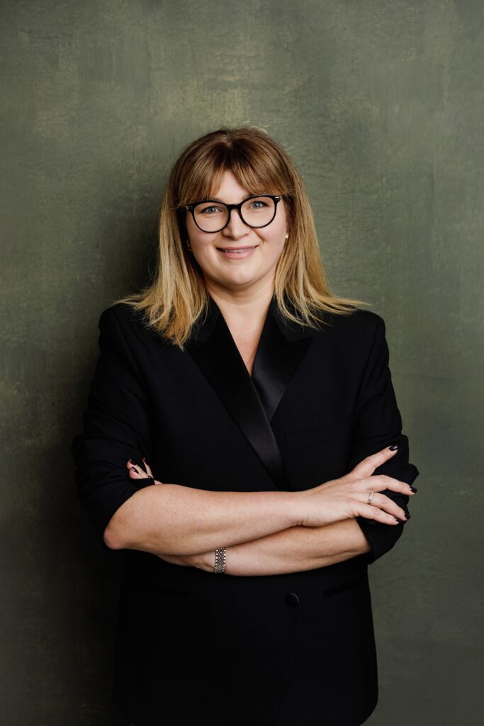 Professional headshot of a woman with glasses and a blonde fringe in a black blazer, arms folded, smiling softly against a green textured backdrop