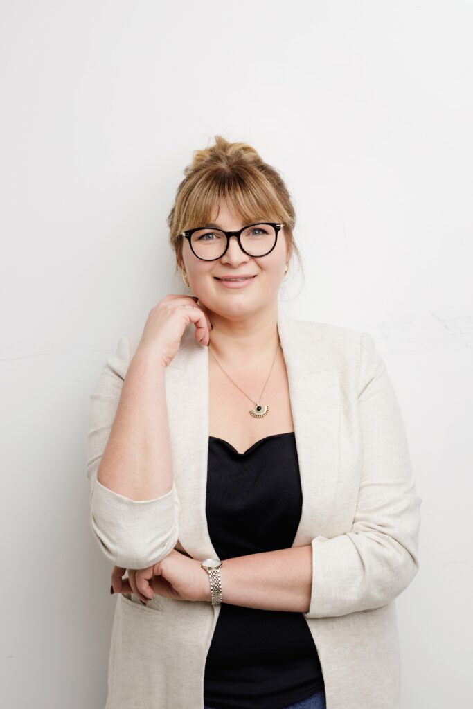 Bright headshot of a woman with glasses in a cream blazer, resting her chin on her hand against a white backdrop