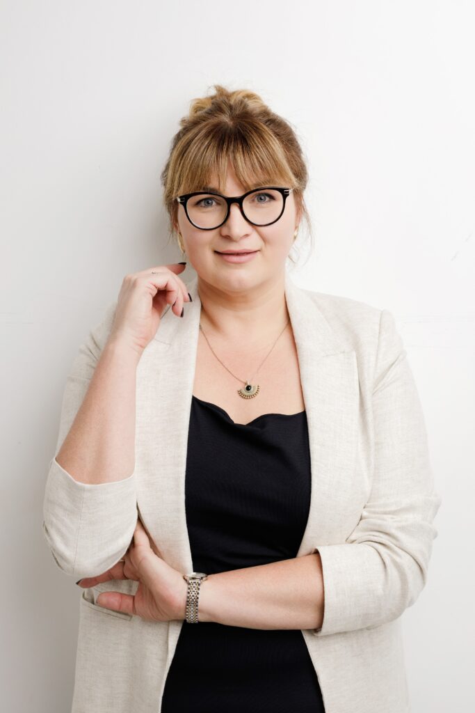 Relaxed professional headshot of a woman with glasses and her hair up in a cream blazer, arms folded, against a white backdrop